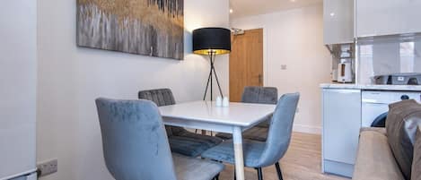 Un comedor moderno con una mesa blanca y sillas tapizadas grises, una lámpara de pie negra, y una cocina al fondo.