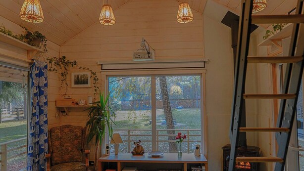 A cozy room with a wooden ceiling, a large window with a view of trees, a bookshelf, a chair, and a small table with a teddy bear and a vase.