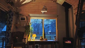 A cozy room with a wooden ceiling, a large window, a bookshelf, a lamp, and a heater. - Metai Miške&a Year in the Forest (Paliepis)