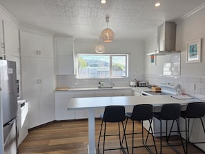 A modern kitchen with white cabinets, a central island with bar stools, a window with blinds, and a hanging light fixture. - Seascape Retreat, breathtaking sea views. (Riverton / Aparima)