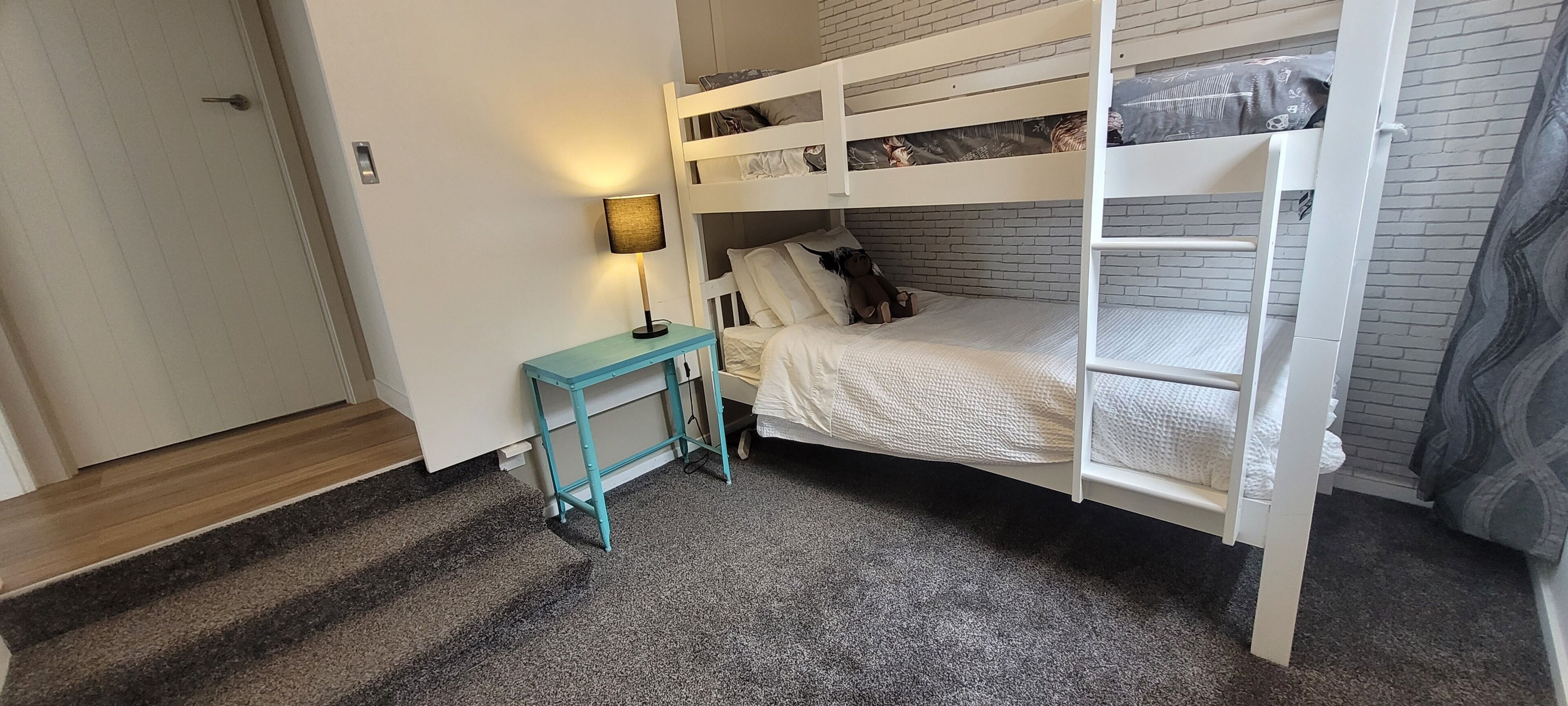 A bunk bed room with a small table and lamp, carpeted floor, and a door.