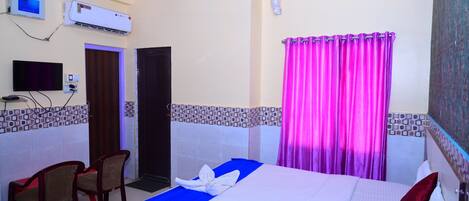 Habitación de hotel con cama, dos sillas, aire acondicionado, ventilador de techo, televisor y puerta de baño.