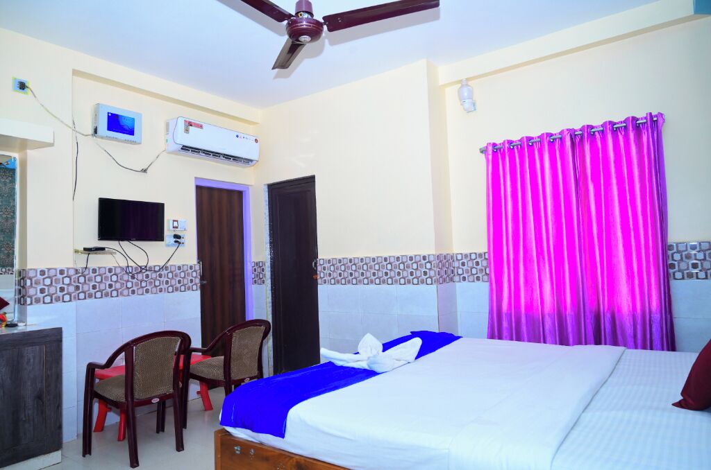 A hotel room with a bed, two chairs, a desk, an air conditioner, a television, and a ceiling fan.