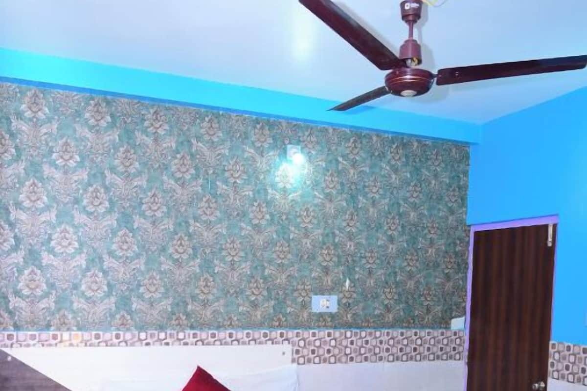 Una cama bien tendida con una colcha azul y ropa blanca, un ventilador de techo y papel tapiz estampado.