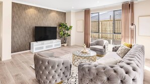 A modern living room with a flat-screen TV, a sofa, armchairs, a coffee table, and a large window with curtains.
