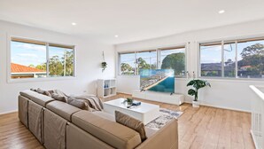 Ein modernes Wohnzimmer mit einem beigefarbenen Ecksofa, einem weißen Couchtisch und einem großen Fernseher, der eine Küstenlandschaft zeigt.
