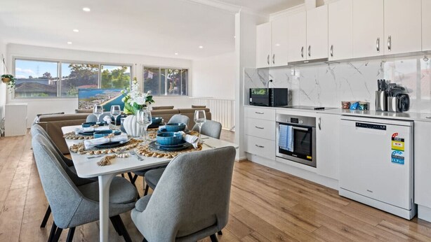 Eine moderne Küche mit weißen Schränken, einer Mikrowelle, einem Backofen und einem Geschirrspüler. Ein Essbereich mit Tisch für zwei Personen, Stühlen und Blick auf eine malerische Landschaft durch große Fenster.