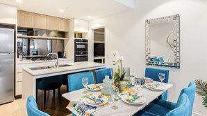 Una cocina moderna con una isla central, electrodomésticos integrados y un comedor con sillas azules y una mesa blanca para seis personas.