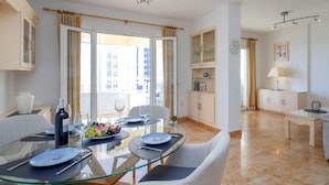 A modern dining area with a glass table, white upholstered chairs, and a view of the city through large windows. - Apartment Oasis by Interhome (Calpe/Calp)