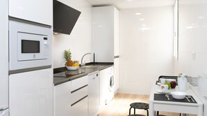 A modern kitchen with white cabinetry, a microwave, a sink, and a washing machine. There is a dining area with a table, chairs, and a small island with a bowl and spoon.