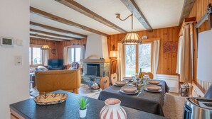 A rustic cabin interior with a dining area, wooden beams, a fireplace, and a television in the living room.