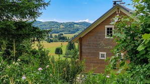 A wooden cabin with a gabled roof, surrounded by lush greenery and fields.