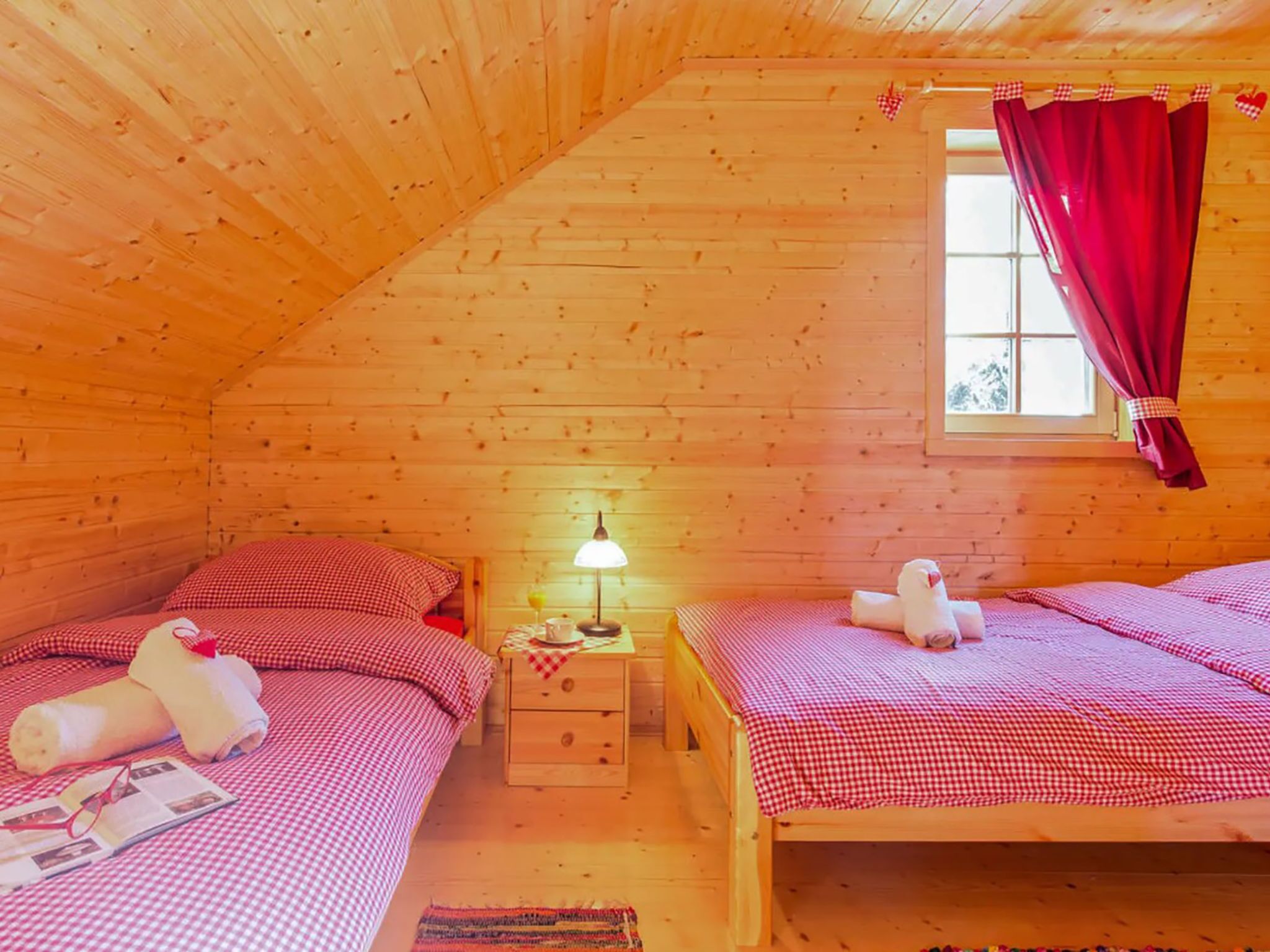 Ein gemütliches Schlafzimmer einer Holzhütte mit zwei Betten, einem Fenster mit roten Vorhängen, einem kleinen Nachttisch mit einer Lampe und einem Stofftier auf einem Bett.