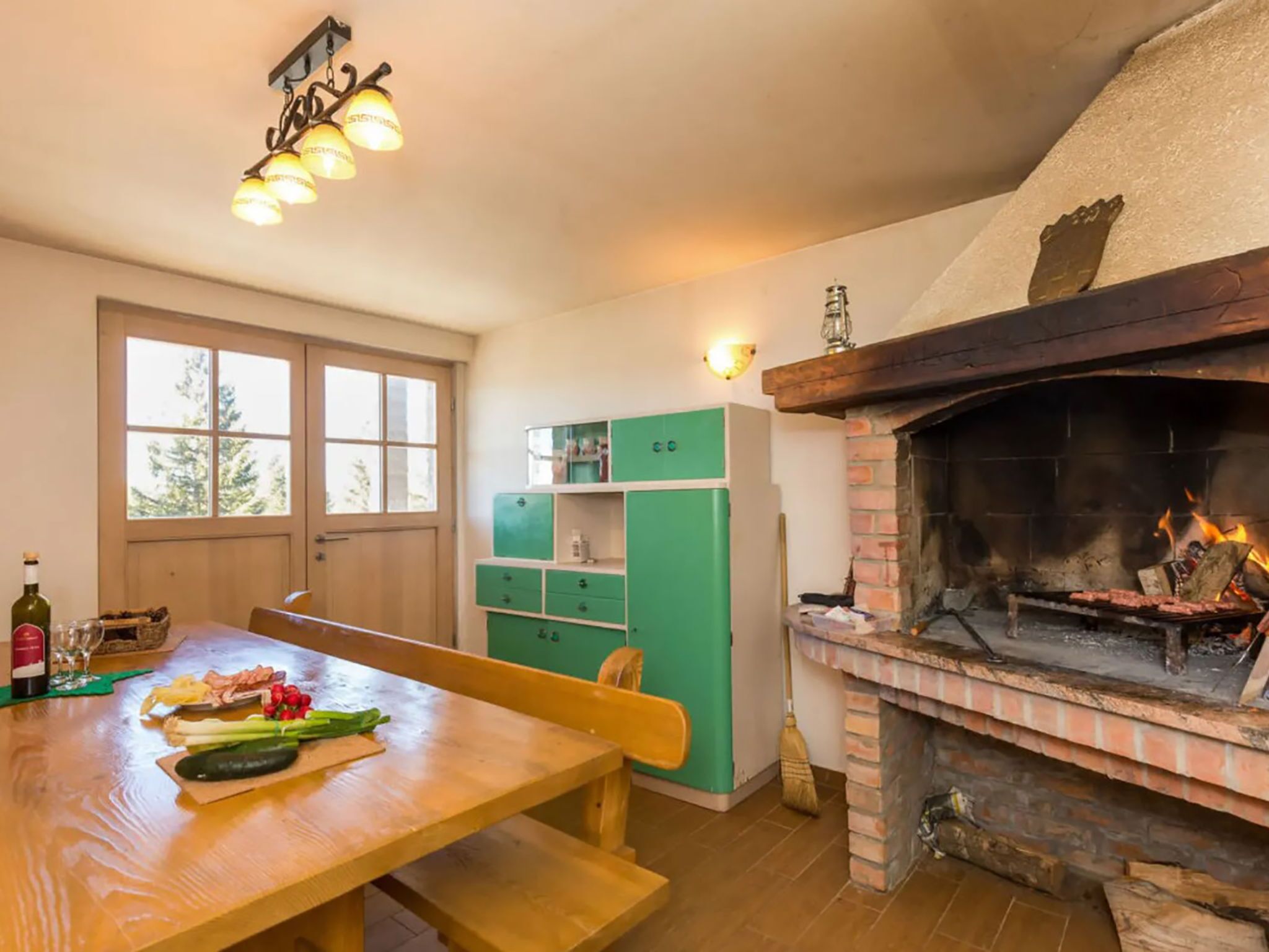 A cozy kitchen with a brick fireplace, wooden table, and a window with a view of trees.