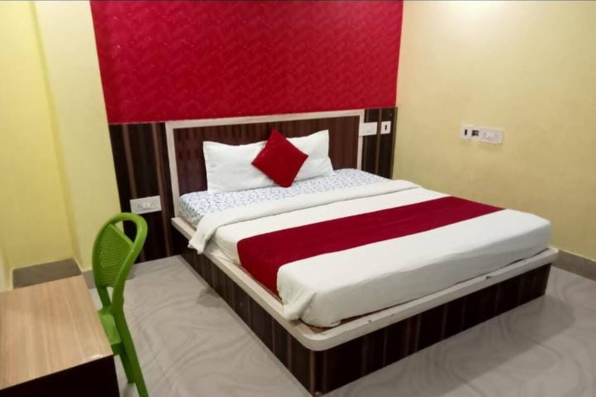 Un lit simple avec un oreiller rouge, une couverture blanche et un plaid rouge. Une chaise verte se trouve à gauche du lit.
