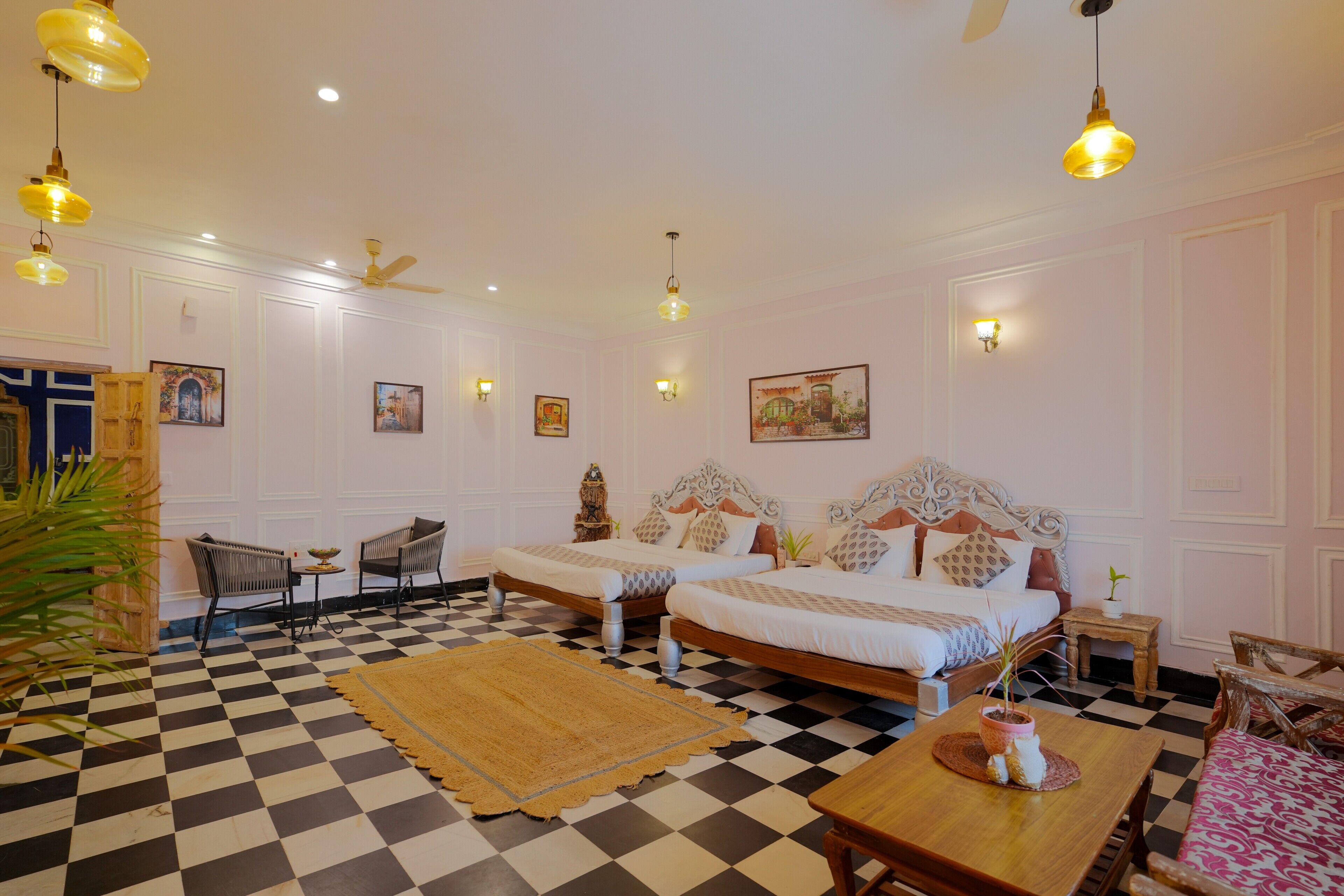 A hotel room with a checkered floor, two beds with ornate headboards, a dining table, and a ceiling fan.