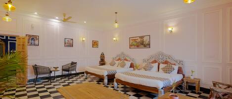 A hotel room with a checkered floor, two beds with ornate headboards, a dining table, and a ceiling fan.