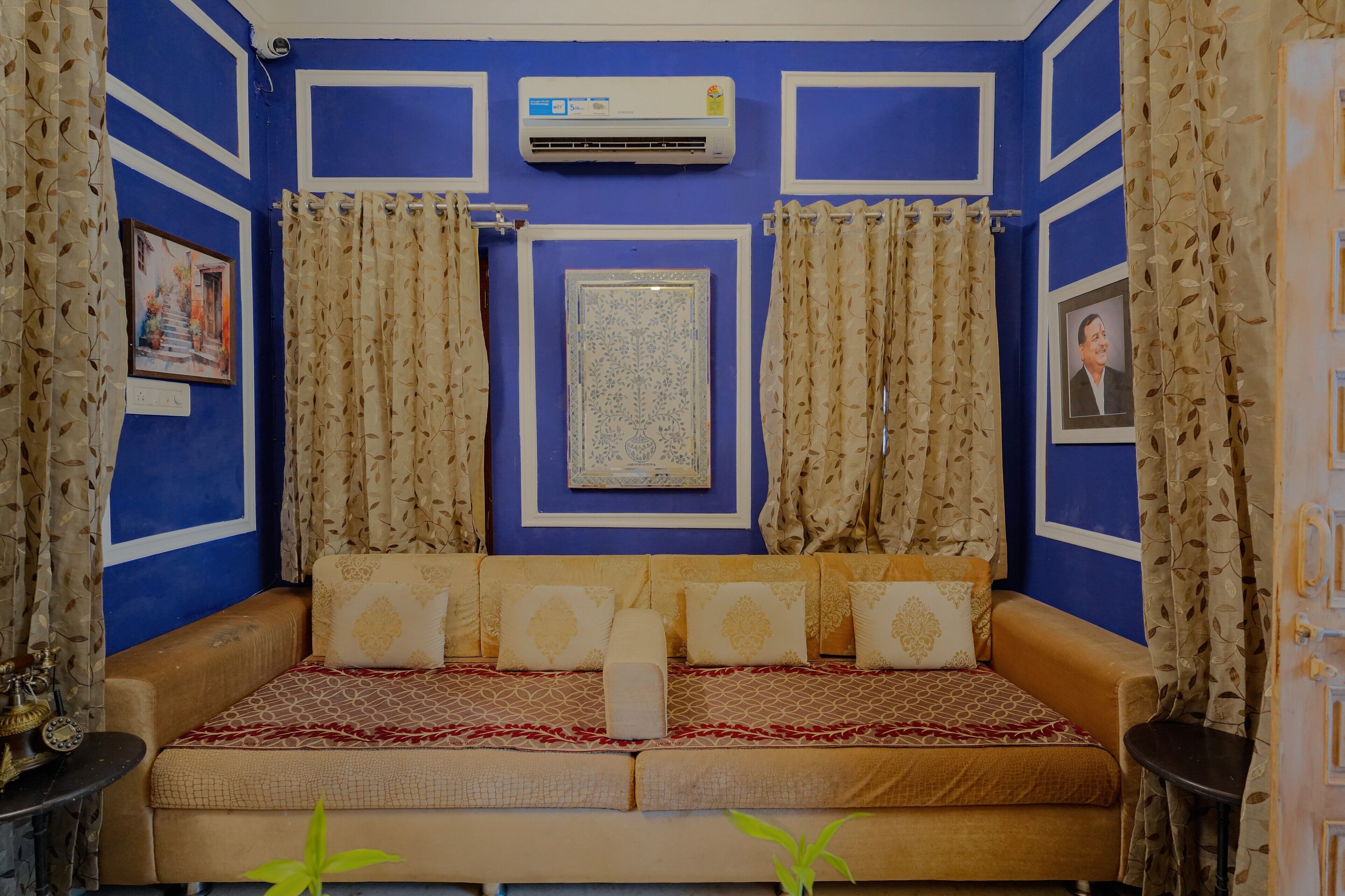 A room with a blue wall, a sofa with patterned cushions, a framed picture, and an air conditioning unit.