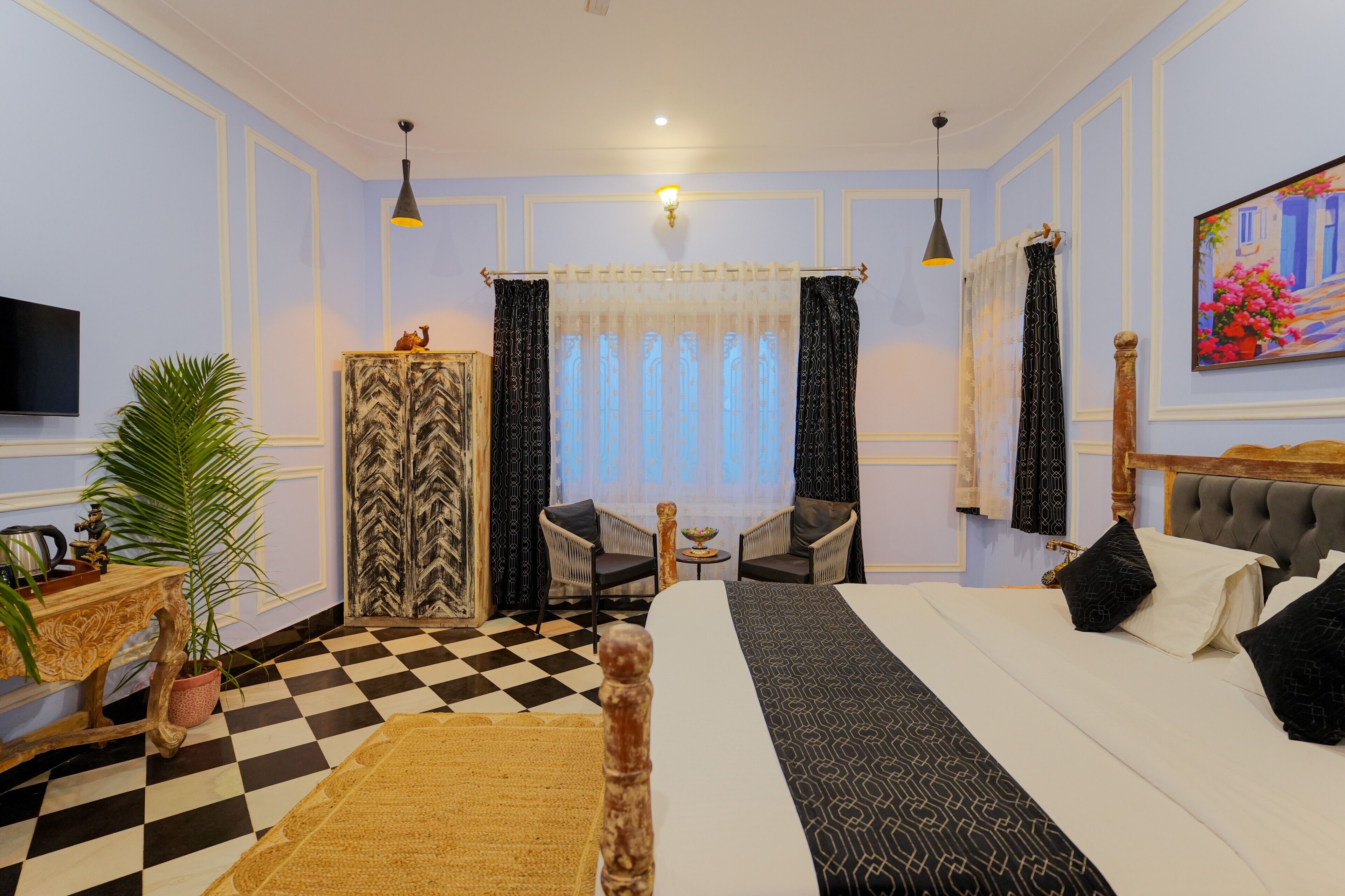 A hotel room with a checkered floor, a bed with patterned bedding, two chairs, a small table with a bowl, a potted plant, and a painting on the wall.