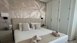 Un lit bien fait, avec deux serviettes enroulées, une tête de lit ornée d’un motif de feuilles de palmier et une table de chevet avec une lampe et une petite plante.