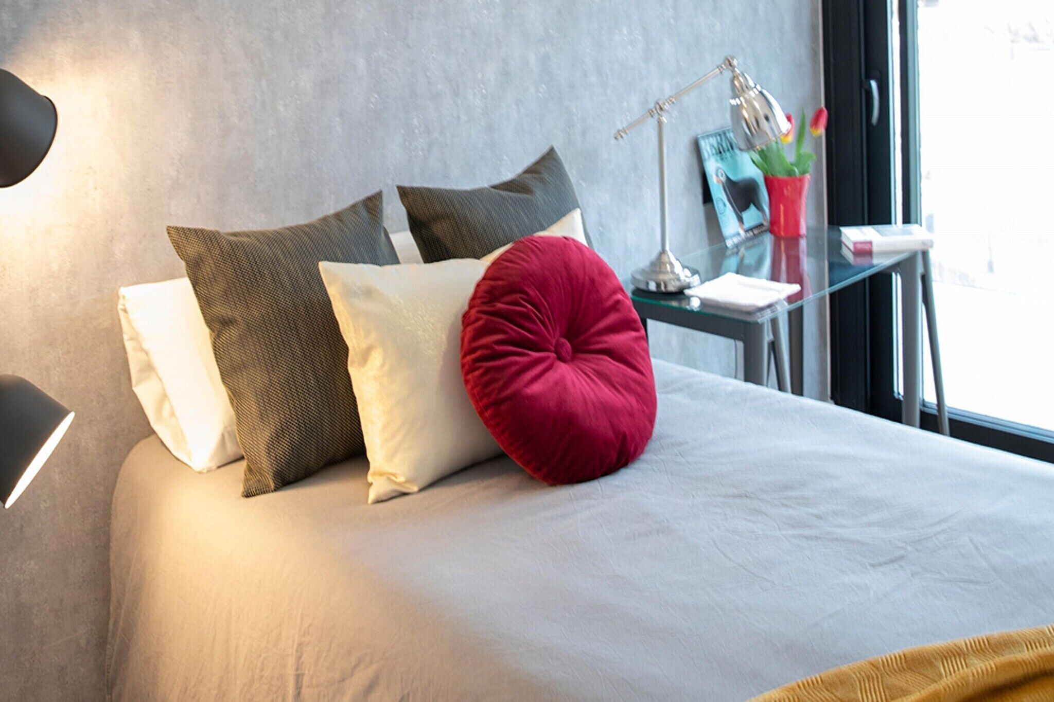 Een opgemaakt bed met een wit en rood kussen, een lamp en een tafeltje met een plant en een boek.