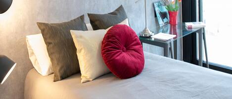 Een opgemaakt bed met een wit en rood kussen, een lamp en een tafeltje met een plant en een boek.