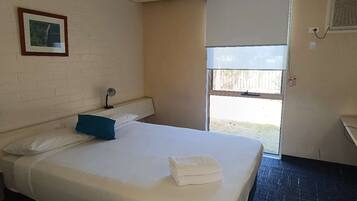 A single-bed room with a white bed, a blue pillow, a white towel, a picture on the wall, and a window with a view of a fence.