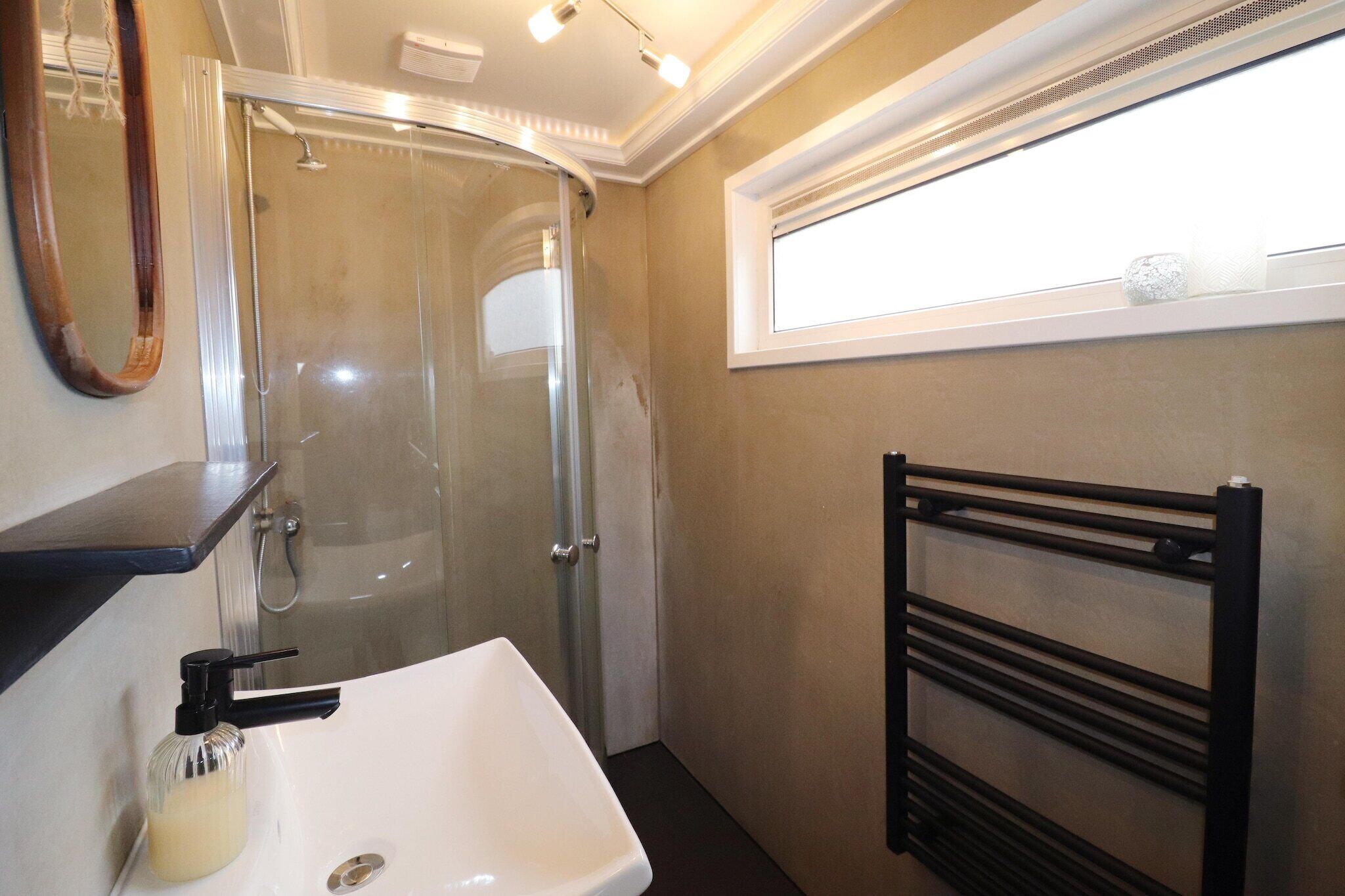 A bathroom with a glass shower enclosure, a white sink, a black towel rack, and a window above the sink.