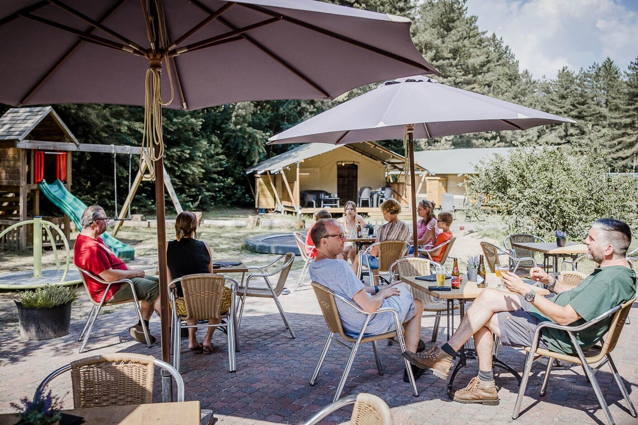 Un espacio al aire libre con un patio que tiene sillas y mesas de metal, sombrillas y un parque infantil al fondo.