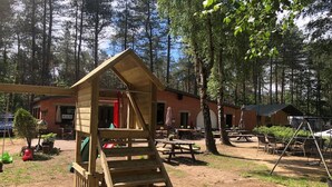 A wooden play structure in a forested area with picnic tables and a brick building in the background.