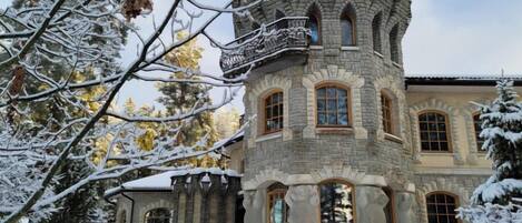 A stone building with multiple windows and a balcony, surrounded by snow-covered trees.