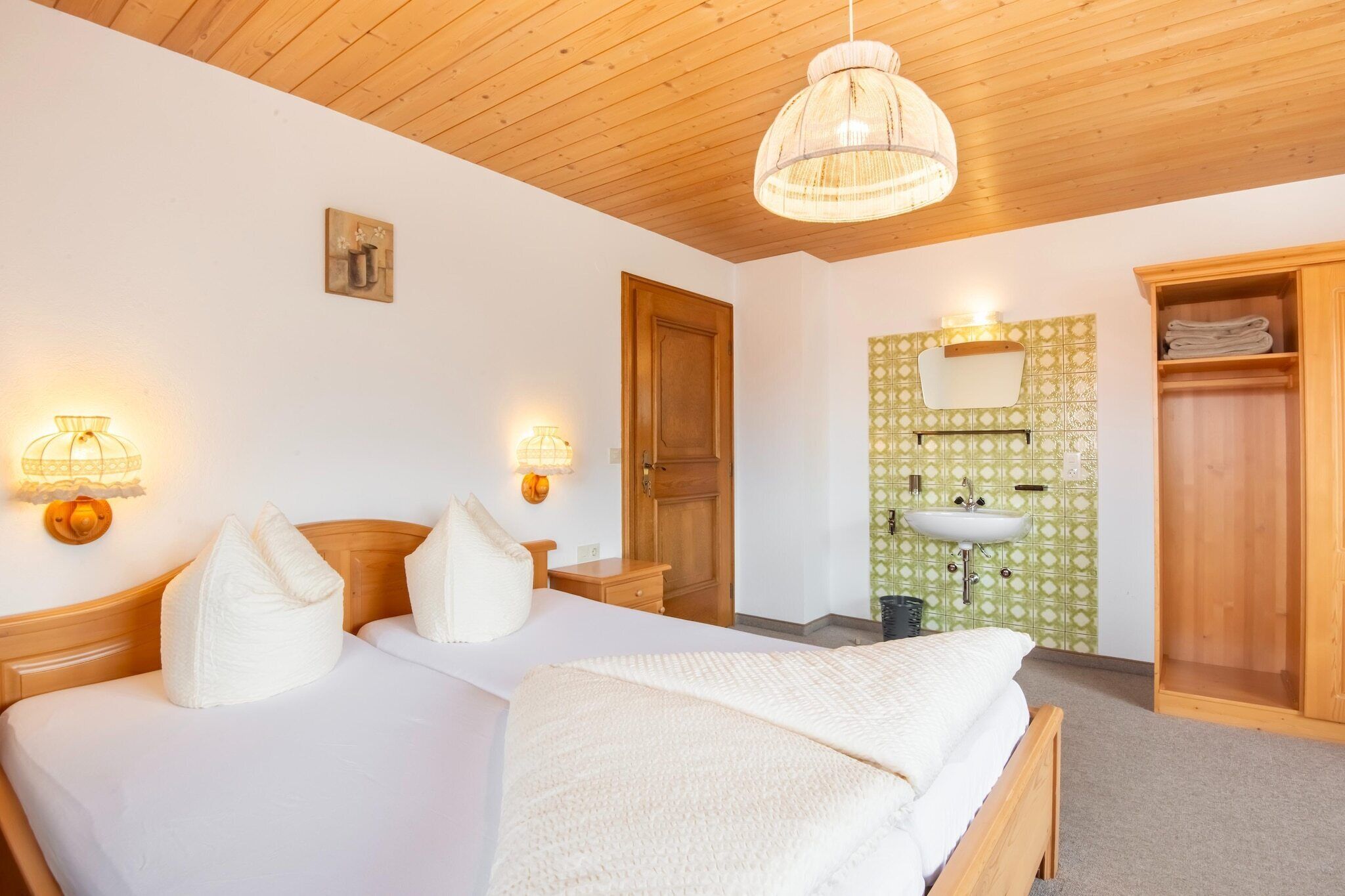 A bedroom with a wooden ceiling, two bedside tables with lamps, a bed with white bedding, a wooden wardrobe, and a green patterned wall.