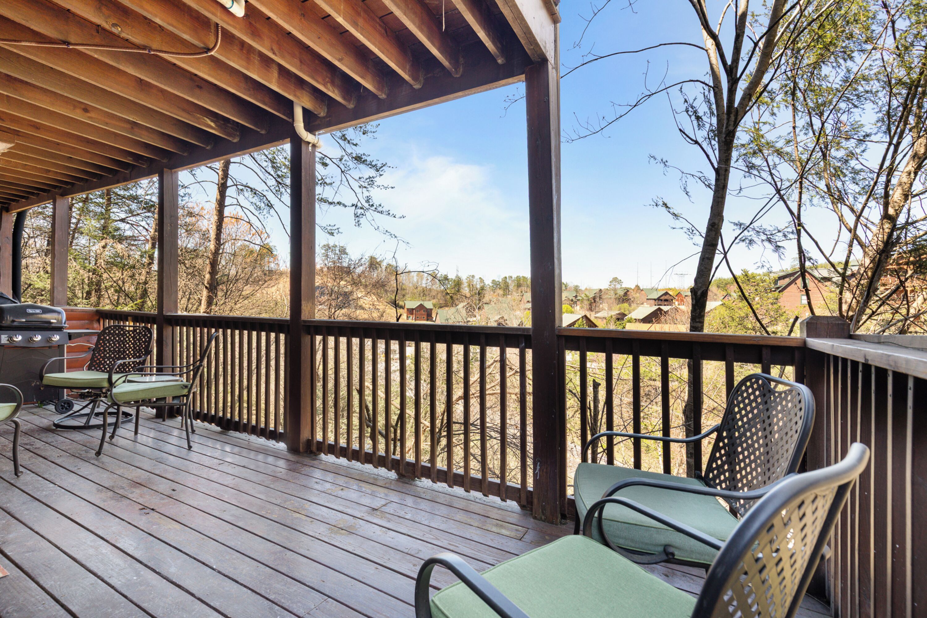 A covered deck with wooden railings, green cushioned chairs, and a grill.