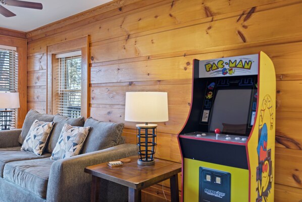 A room with a wooden wall, a couch with patterned pillows, a lamp, and a Pac-Man arcade machine.