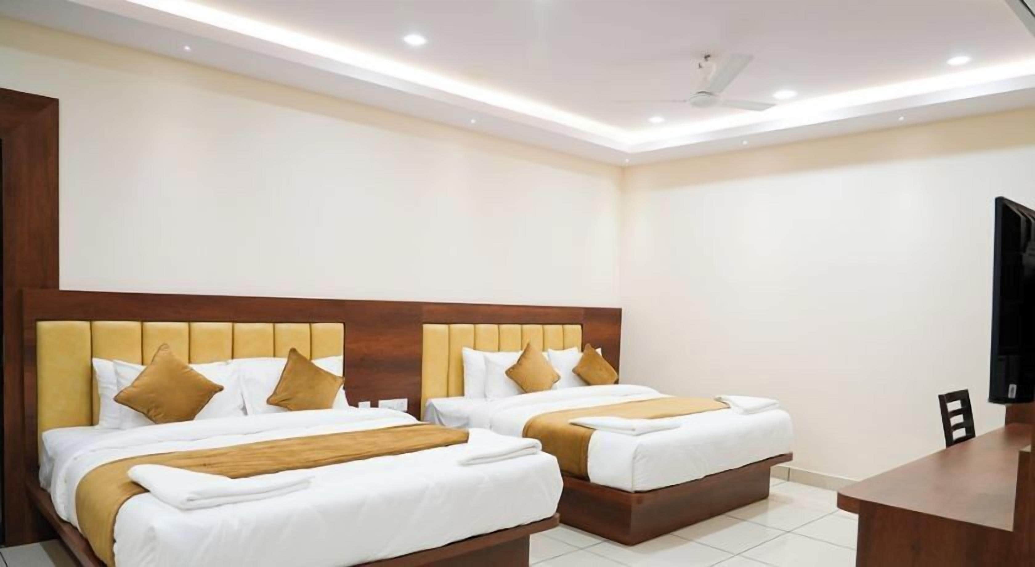 A hotel room with two beds, each with a wooden headboard and white bedding. There is a flat-screen TV on a wooden stand to the right.