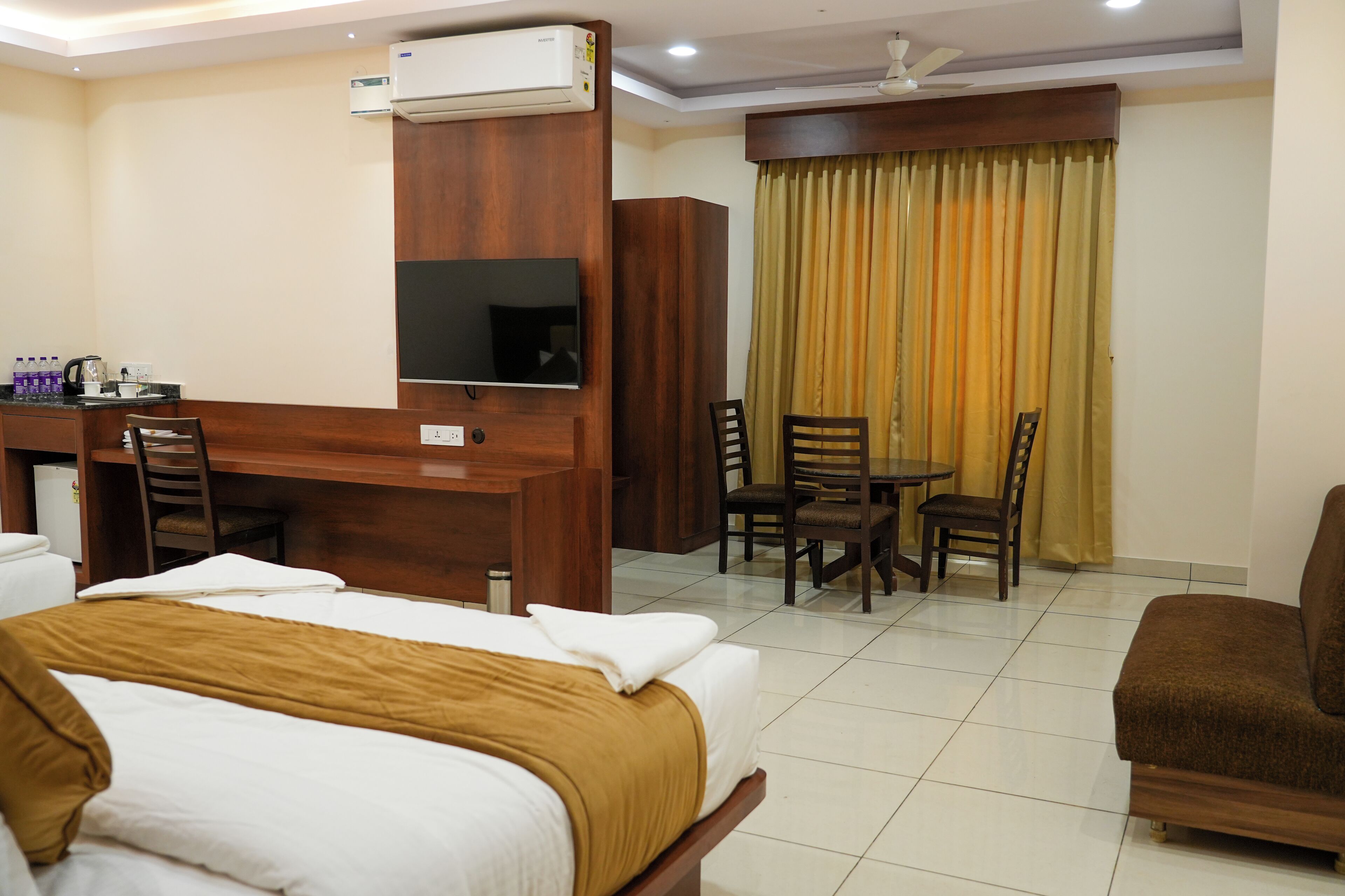 A hotel room with a bed, a desk with a chair, a TV, and a dining area with chairs.