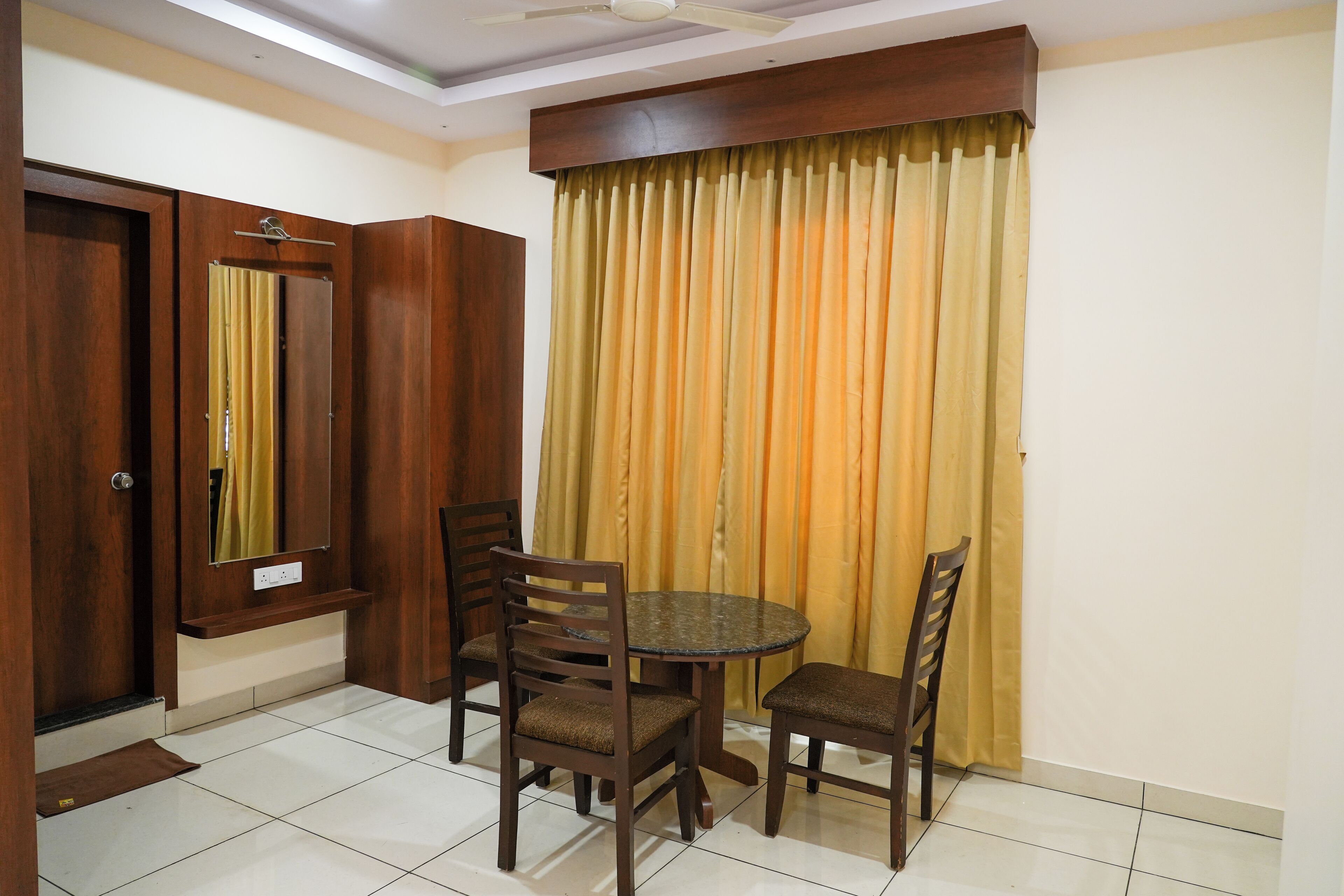 Um quarto com uma mesa redonda, quatro cadeiras, um guarda-roupa de madeira, um espelho e uma cortina.