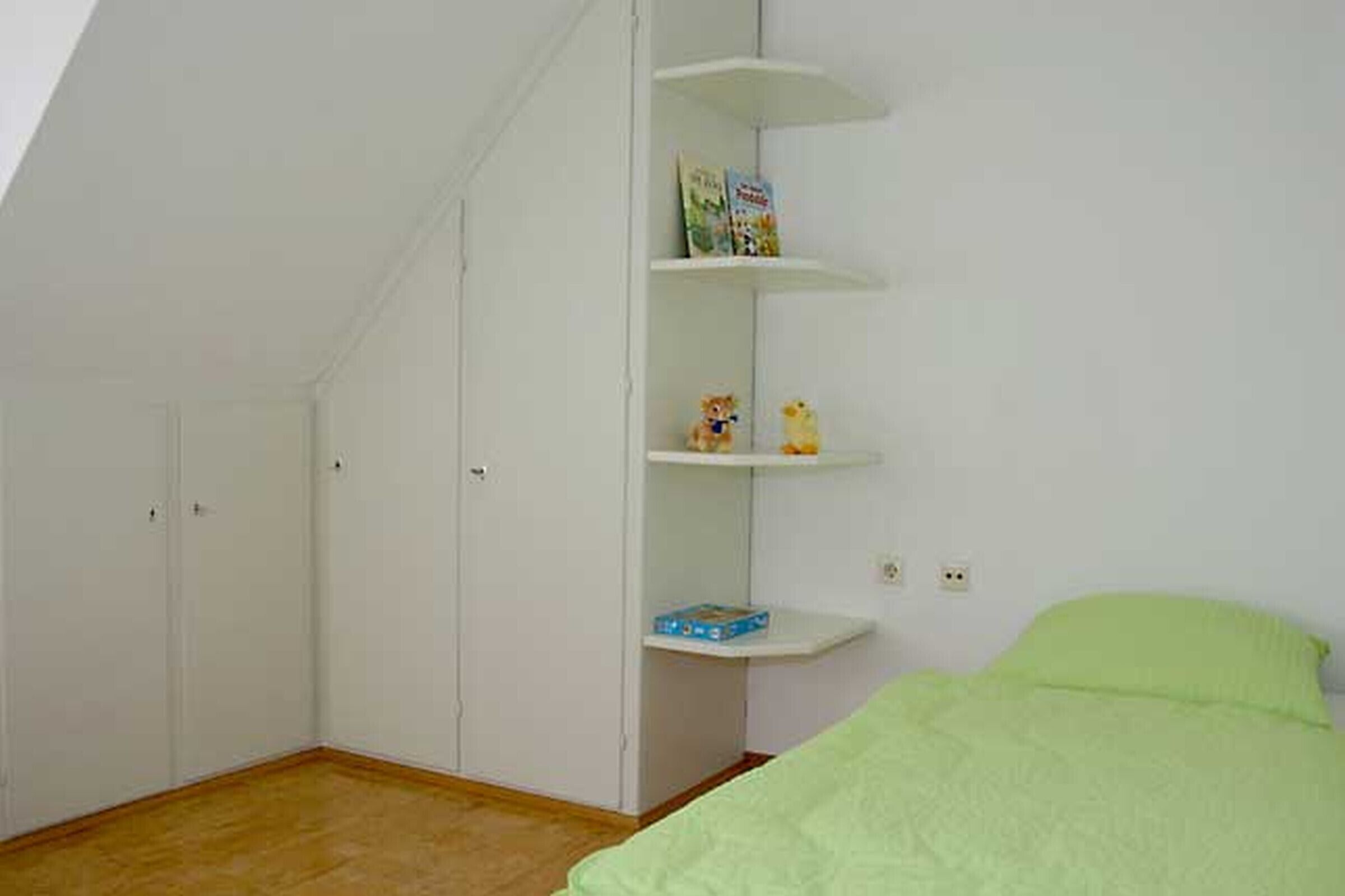 A bedroom with a sloped ceiling, a bed with a green cover, white walls, and a built-in shelving unit with books and figurines.