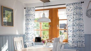 Un coin repas avec une table en bois, des chaises blanches, une suspension et une fenêtre agrémentée de rideaux à motifs floraux.