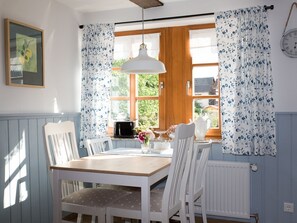Un comedor con una mesa de madera, sillas blancas, una lámpara colgante y una ventana con cortinas florales.