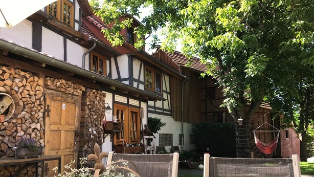 Un bâtiment traditionnel à colombages, avec une porte en bois, un mur de pierre où est empilé du bois de chauffage et une chaise suspendue.