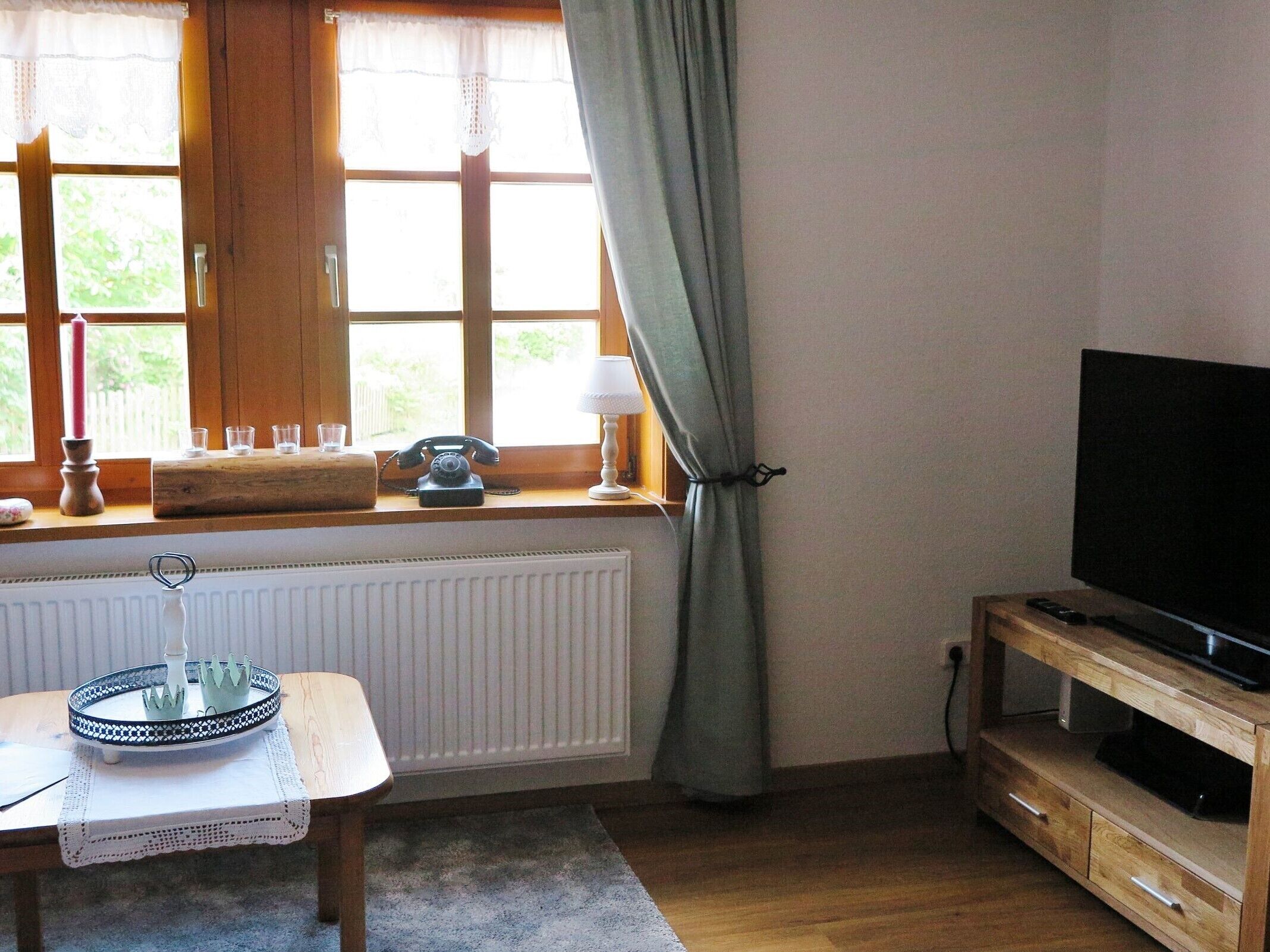 Une pièce avec une table en bois, un téléviseur posé sur un meuble en bois, une fenêtre avec des rideaux et un radiateur.
