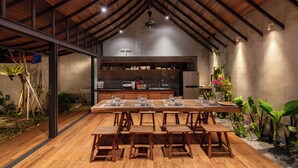 A spacious dining area with a long wooden table and benches, a kitchen in the background, and a high ceiling with exposed beams. - AnNam Village 1 (Vung Tàu)