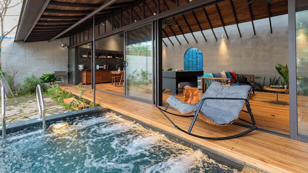A modern indoor pool area with a wooden deck, a lounge chair, and a dining area with a blue window. - AnNam Village 1 (Vung Tàu)