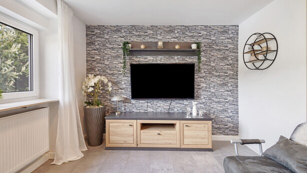 Un salon moderne avec un mur en pierre, un téléviseur fixé au mur, une console en bois et une fenêtre avec des rideaux.