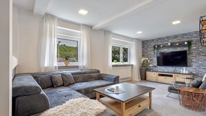 Un salon moderne comprenant un canapé d’angle, une table basse, un téléviseur fixé à un mur en pierre et un coin repas avec une table et des chaises en bois.