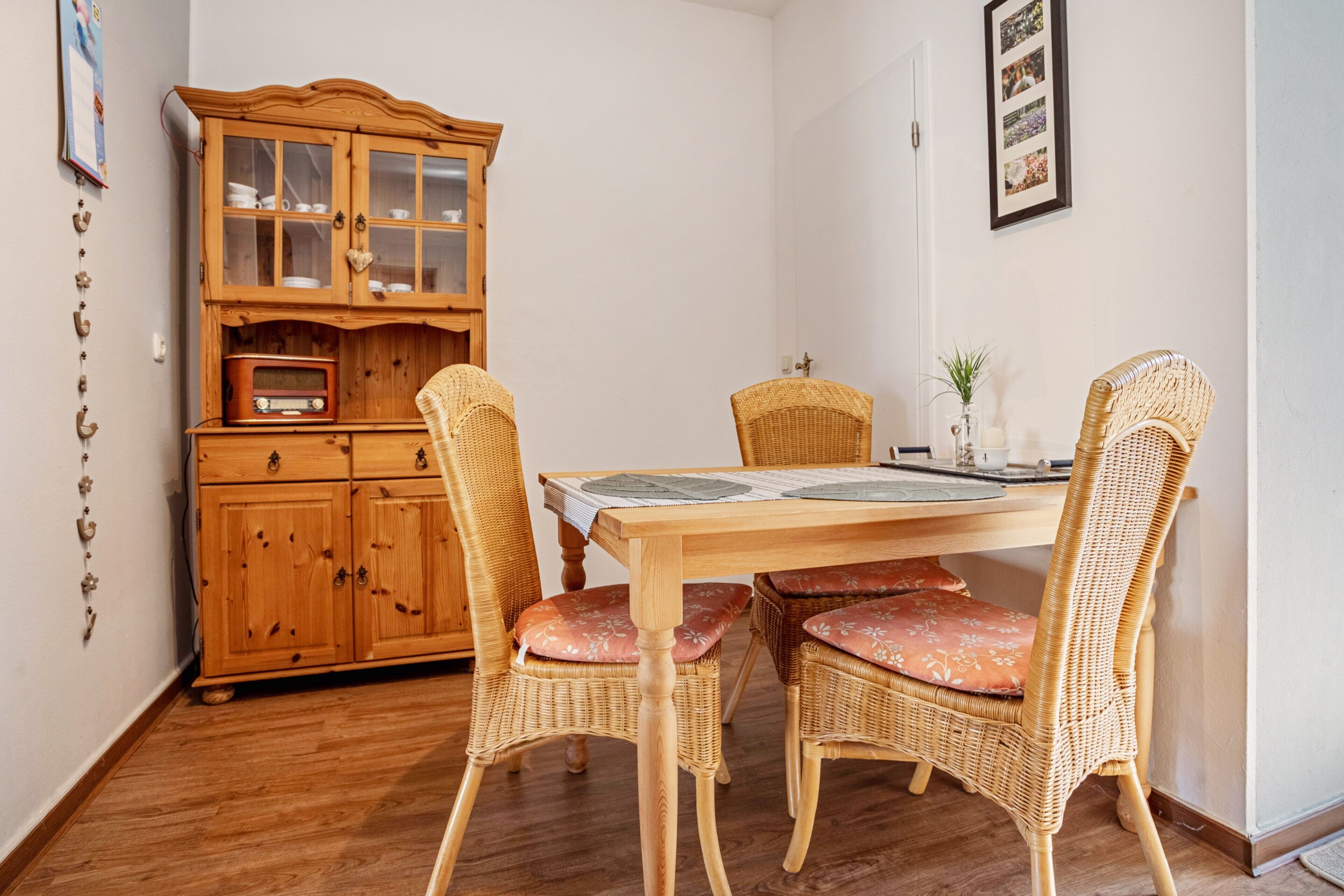 Un coin repas avec une table en bois, des chaises en osier ornées de coussins à motifs floraux, un buffet en bois doté de portes en verre et une radio d’époque.