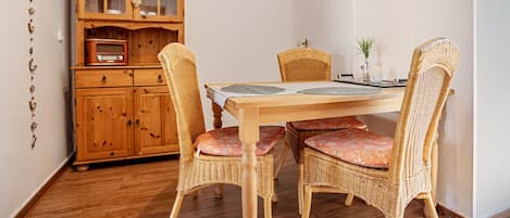 Un coin repas avec une table en bois, des chaises en osier ornées de coussins à motifs floraux, un buffet en bois doté de portes en verre et une radio d’époque.