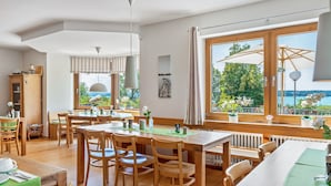 Ein Essbereich mit Holztischen und -stühlen, Blick auf das Meer und ein großes Fenster mit Vorhängen.