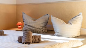 Un letto rifatto con due cuscini, una lampada rossa e un asciugamano arrotolato su una parete beige.